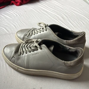 Grå sneakers från Axel Arigato - Snygga grå sneakers från Axel Arigato i skinn med mockadetalj på hälen. Klassisk låg modell med rund tå, vita snören och platt sula. Perfekta för en clean och stilren look.