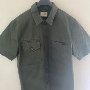 Grön overshirt från Nudie Jeans -  Stilren grön overshirt från Nudie Jeans med korta ärmar och klassisk krage. Skjortan har två bröstfickor, varav en med knapp, och stängs med knappar framtill. Tillverkad i bomull och passar perfekt för en avslappnad look.