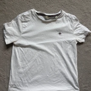 Vit t-shirt från GANT XS - Klassisk vit t-shirt från GANT i storlek XS. T-shirten har rund halsringning, korta ärmar och en broderad GANT-logga med sköld på bröstet. Tillverkad i mjuk bomull för en skön känsla. Perfekt basic-plagg med stilren design.
