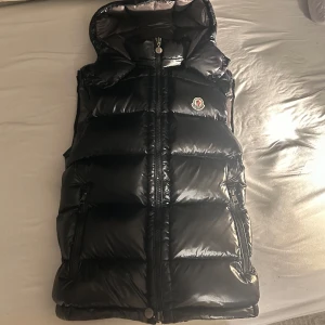 Moncler väst - Snygg svart dunväst från Moncler med glansig finish och huva. Västen har dragkedja framtill, två fickor med dragkedja och klassisk Moncler-logga på bröstet. Insidan har coola etiketter och serier. Perfekt för lager-på-lager-stil under kyliga dagar.