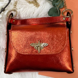 Metallic orange axelväska med guldkedja skinn Italiensk Bee bag Collection - Cool handgjord Italiensk axelväska  i skinn metallic orange med guldfärgade detaljer och kedja. Från Bee bag Collection. Framsidan har en snygg guldfärgad insekt som dekoration. Väskan är boxig i formen och har tryckknappar på sidorna. Perfekt för dig som vill sticka ut med accessoarer. Denna dubbla handtagsväska har en avtagbar rem som gör att du kan bära den bekvämt på axeln eller som en remväska.  Äkta läder och handgjort i Italien. Färg: orange metallic  Mått: 19 cm x 14 cm x 6 cm
