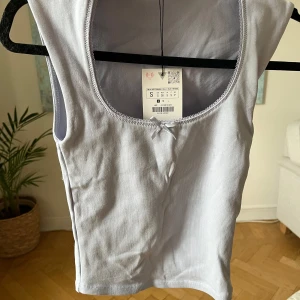 Ljuslila linnetopp med rosett - Supersöt ljuslila linnetopp med rundad urringning och liten rosett framtill. Toppen är ärmlös och har en fin, diskret spetskant längs halsringningen. Perfekt att styla med jeans eller kjol för en chill look.