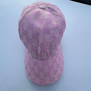 Rosa Gucci keps med GG-mönster - Helt oanvänd och snygg rosa keps från Gucci med ikoniskt GG-mönster över hela kepsen. Justerbar rem baktill och böjd skärm. Tillverkad i slitstark bomullsmix med broderade detaljer. Perfekt statement-piece för dig som gillar lyxiga accessoarer.