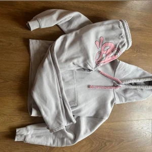Syna World tracksuit - Säljer en ljusgrå tracksuit från Syna World med rosa snören och detaljer. Tröjan har huva och magficka, perfekt för en chill och avslappnad stil. Mjuk och skön!
