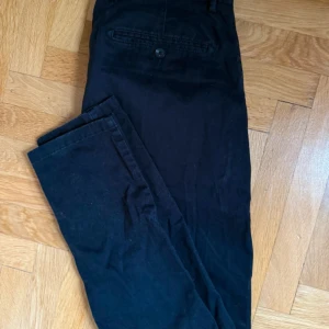 Svarta chinos från John Henric W31 - Svarta chinos från John Henric i klassisk rak modell. Byxorna har sidfickor, bakficka med knapp och bälteshällor. Tillverkade i bomull med en stilren look som funkar till många outfits.