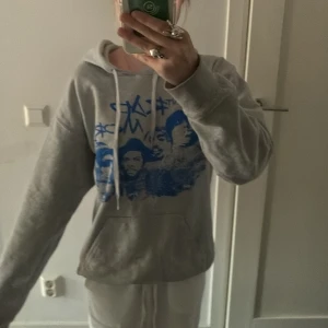 Grå hoodie med blått tryck - Säljer en grå hoodie med stort blått grafiskt tryck framtill och vita snören i huvan. Tröjan har känguruficka och en avslappnad passform, perfekt för chill dagar. Motiv med text och ansikten i blått ger en cool streetkänsla.