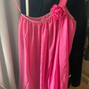 Rosa kortklänning med blomdetalj Bik Bok - Supersöt rosa kortklänning från Bik Bok med tunna axelband och en stor tygblomma på ena axelbandet. Klänningen har ett luftigt, veckat tyg och en lätt A-linjeform. Perfekt för dig som vill sticka ut med en färgstark look!