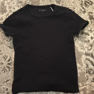 Svart ribbad t-shirt från Reserved - En stilren svart t-shirt från Reserved i ribbat material. Klassisk rund halsringning och korta ärmar. Perfekt basplagg med en clean look som funkar till allt. Skön passform och enkel att matcha med dina favoritjeans eller kjol.