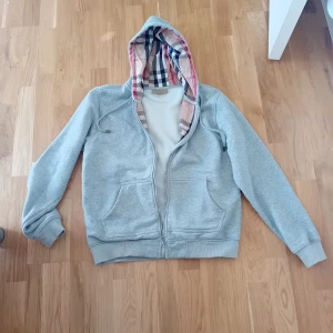 Grå hoodie från Burberry med dragkedja - Säljer en grå hoodie från Burberry med klassiskt rutigt foder i huvan. Tröjan har dragkedja framtill, två fickor och ribbade muddar. Perfekt för dig som gillar stilrena och bekväma plagg med en touch av lyx.