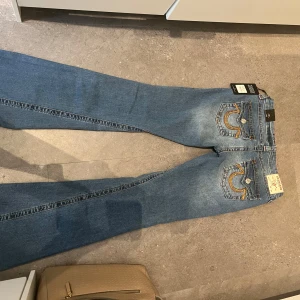 True religon bootcut jeans blå med broderi - Snygga blå bootcut jeans från true religon med coola broderade detaljer på bakfickorna och slitningar framtill. Jeansen har klassisk femficksdesign, orangea sömmar och gylf med dragkedja. Perfekta för dig som gillar en retro vibe och vill sticka ut lappen e kvar och är helt nya !! 