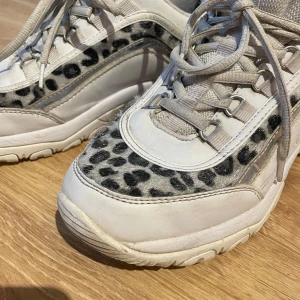 Fila sneakers med leopardmönster Fila - Chunky vita Fila sneakers med coola leopardmönstrade detaljer och silvriga inslag på sidorna. Skorna har snörning, platt sula och rund tå. Ovandelen är i syntet och textil, vilket ger en sportig och trendig vibe. Perfekta för dig som vill sticka ut med din stil.