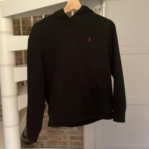 Svart hoodie från Polo Ralph Lauren - Svart hoodie från Polo Ralph Lauren med klassisk röd broderad logga på bröstet. Tröjan har huva, känguruficka och långa ärmar. Perfekt för en chill och stilren look. Materialet känns mjukt och bekvämt mot huden.
