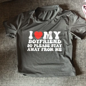 Svart croppad topp med texttryck - Svart croppad topp i polyester med vit och röd text: 'I ❤️ MY BOYFRIEND SO PLEASE STAY AWAY FROM ME'. T-shirten har korta ärmar och en tight passform, perfekt för dig som vill sticka ut med ett statement-plagg.