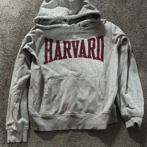 Grå Harvard hoodie med huva - Snygg grå hoodie med stor vinröd HARVARD-text på bröstet. Tröjan har huva med snörning och en stor magficka framtill. Perfekt för dig som gillar college-vibe och vill ha en bekväm och stilren look.