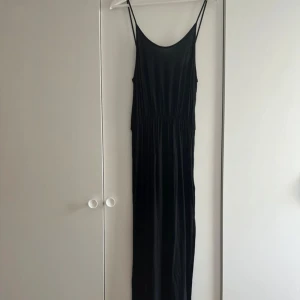 Svart långklänning med smala axelband - En svart långklänning med smala axelband och rundad urringning. Klänningen har en avslappnad passform med resår i midjan som ger en snygg siluett. Tillverkad i ett mjukt, tunt material som faller fint