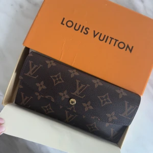 Louis Vuitton Monogram Plånbok - Snygg plånbok från Louis Vuitton i klassiskt monogrammönster. Mörkbrun med ljusbruna LV-loggor och blommor, tillverkad i tåligt skinn. Flera kortfack, fönster för ID och dragkedjefack för mynt. Lyxig och tidlös accessoar med guldknapp.