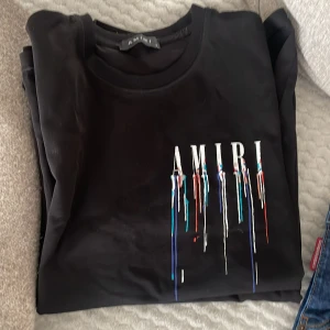 Svart AMIRI t-shirt med färgstänk - Svart t-shirt från AMIRI med stort vitt logotryck framtill och coola färgstänk i blått, rött och vitt som rinner ner från bokstäverna. Klassisk rund hals och korta ärmar. Tillverkad i mjuk bomull för skön passform.