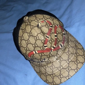 Gucci keps med orm och GG-mönster - Snygg keps från Gucci med klassiskt GG-monogram i beige och brunt, dekorerad med en röd och vit orm på framsidan. Baksidan är i svart mesh och justerbar med grön-röd randig rem. Perfekt statement-piece för dig som vill sticka ut. För bara 700 MALMÖ!!