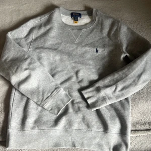 Grå sweatshirt från Polo Ralph Lauren - Klassisk grå sweatshirt från Polo Ralph Lauren med rund halsringning och broderad marinblå logga på bröstet. Tröjan har långa ärmar och ribbade muddar vid ärmslut och nederkant. Perfekt för en chill och stilren look.