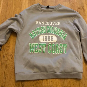 Grå sweatshirt med Vancouver-tryck - Grå sweatshirt med tryck i vitt och grönt där det står 'Vancouver British Columbia 1886 West Coast' på framsidan. Tröjan har rund halsringning och långa ärmar. Perfekt för en avslappnad och cool stil.
