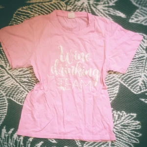 Rosa t-shirt med texttryck - Ljusrosa t-shirt med vit text 'Wine drinking TEAM' på bröstet. Klassisk rund halsringning och korta ärmar. Skön och avslappnad passform, perfekt för chill dagar. Tillverkad i mjuk bomull för en bekväm känsla.