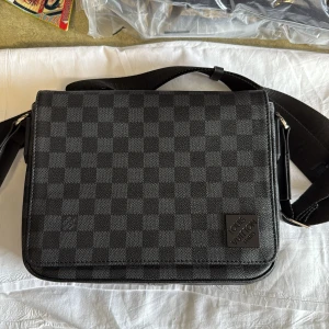 Louis Vuitton axelväska Damier Graphite - Snygg axelväska från Louis Vuitton i klassiska Damier Graphite-mönstret. Väskan är svart och grå med rutigt mönster, justerbar axelrem med logga och dragkedja baktill. Tillverkad i tåligt skinn och textil, perfekt för dig som gillar stilren design.