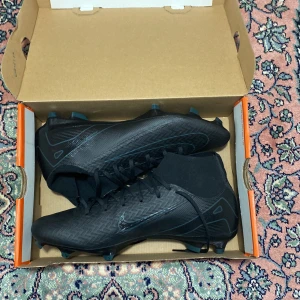 Svarta Nike fotbollsskor med snörning - Snygga helsvarta Nike fotbollsskor med hög strumpliknande krage och snörning. Skorna har mönstrad ovandel i syntetmaterial och fasta dobbar för gräsplan. Perfekta för dig som vill ha en clean look på planen.