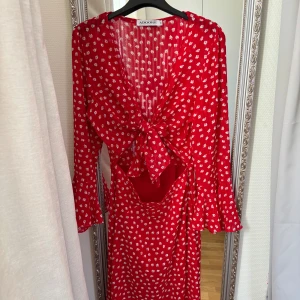 Röd blommig klänning från Adoore - Supersöt röd klänning från Adoore med vita blommor, långärmad med volang i ärmslut och stor knytrosett framtill. Djup v-ringning och cut-out vid midjan ger en trendig vibe. Perfekt för sommarens alla tillfällen! Använd en gång, säljs pga inte riktigt min färg💞