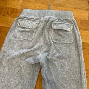 Grå mjukisbyxor Juicy Couture - Grå mjukisbyxor från Juicy Couture med bred resår i midjan och snörning framtill. Byxorna har raka ben, stora fickor både fram och bak samt diskret broderad logga på ena fickan.