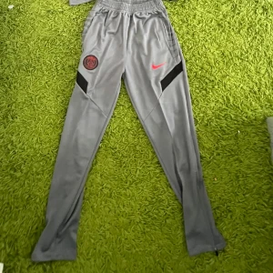 Grå PSG träningsbyxor från Nike - Snygga grå träningsbyxor från Nike med PSG-logga och röd swoosh på låret. Byxorna har smal passform, elastisk midja och dragkedjor vid bensluten. Perfekta för fotbollsträning eller chill.