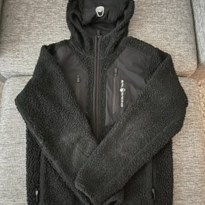 Svart teddyjacka Sail Racing - Svart teddyjacka från Sail Racing med huva och dragkedja. Jackan har mjuk, fluffig yta och detaljer i slät polyester på bröstet med två bröstfickor med dragkedja. Perfekt för kyliga dagar och riktigt skön att bära.