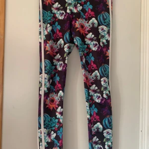 Blommiga leggings från BikBok - Färgglada leggings från BikBok med blommigt mönster i lila, turkos, rosa och vitt. De har en svart bas och vita ränder längs sidorna. Tight passform och stretchigt material som sitter snyggt och bekvämt.