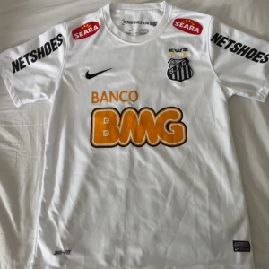 Vit Santos Neymar Jr fotbollströja Nike - Vit Santos FC matchtröja med Neymar Jr och nummer 11 på ryggen. Tröjan har korta ärmar, svarta och orange tryck, klubbmärke på bröstet och flera sponsorer. Tillverkad i Dri-FIT-material som andas. Perfekt för dig som älskar brasiliansk fotboll och klassiska spelare.