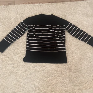 Svartvit randig stickad tröja - Svart stickad tröja med vita horisontella ränder över hela kroppen och ärmarna. Tröjan har lång ärm och en klassisk rund halsringning. Perfekt för dig som gillar en stilren och tidlös look.