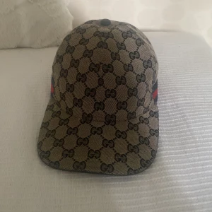 Gucci canvas - Snygg keps från Gucci med klassiskt GG-monogram över hela kepsen i beige och svart. Kepsen har en röd och blå rand på sidan och justerbar rem baktill. Tillverkad i bomullsmix med mesh på insidan för extra komfort. Perfekt för dig som vill sticka ut med en lyxig accessoar.