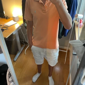 Orange pikétröja från Ralph Lauren - Säljer en snygg orange pikétröja från Ralph Lauren med klassisk krage och knappar framtill. Tröjan har en broderad logga i ljusblått på bröstet. Perfekt för en stilren look!
