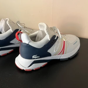 Vita och marinblå sneakers från Lacoste - Snygga sneakers från Lacoste i vitt och marinblått med röda detaljer. Skorna har mesh och syntetmaterial, snörning och rund tå. Klassisk Lacoste-logga på sidan och coola kontrastfärger som ger en sportig vibe.