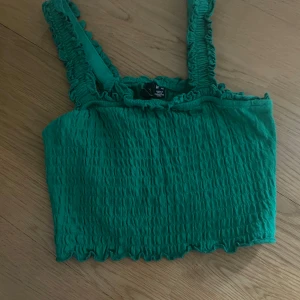 Grön smockad linnetopp Vero Moda XS - Grön smockad linnetopp från Vero Moda i storlek XS. Toppen har breda axelband med volangkant och en croppad passform. Materialet är stretchigt och har en vågig kant nertill för en extra söt look.