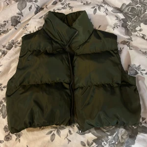 Mörkgrön dunväst med dragkedja - Snygg mörkgrön dunväst med hög krage och quiltad design. Västen har en rak passform och stängs med en svart dragkedja framtill. Perfekt att slänga över en hoodie för en cool och avslappnad look.