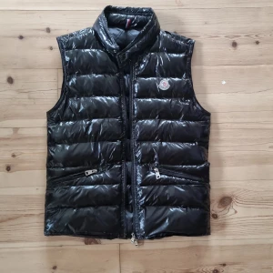 Svart dunväst från Moncler - Snygg svart dunväst från Moncler med glansig finish och klassisk logga på bröstet. Västen har ståkrage, två dragkedjefickor framtill och quiltad design. Perfekt att slänga över en hoodie eller tröja för extra värme.
