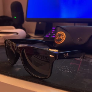 Svarta solglasögon Ray-Ban - Snygga svarta solglasögon från Ray-Ban med klassisk form och logga på skalmen. Tillverkade i blankt syntetmaterial och levereras med originalfodral och putsduk. Perfekta för att lyfta din stil med en ikonisk look.