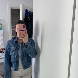 Klassisk blå jeansjacka från Message - Säljer en klassisk blå jeansjacka från Message med bruna knappar och två bröstfickor med lock. Jackan har en figurnära passform och är perfekt att styla till olika outfits. Tidlös modell med snygga söm-detaljer.