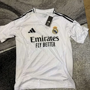 Real Madrid hemmatröja 2023/24 från Adidas - Säljer en vit Real Madrid hemmatröja från Adidas med svarta detaljer och klubbmärke på bröstet. Tröjan har korta ärmar, svart Adidas-logga och tre svarta ränder på axlarna. Materialet är lätt och andas, perfekt för fotbollsträning eller match.