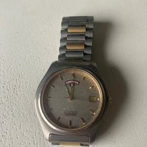 Certina herrklocka i stål och guld - Stilren Certina herrklocka med armband i rostfritt stål och gulddetaljer. Urtavlan är silverfärgad med datumvisning och guldindex. Klassisk design med Certina-logga på spännet. Perfekt för dig som gillar tidlösa accessoarer.