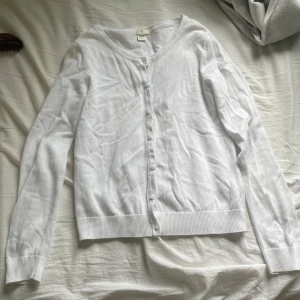 Vit kofta från H&M BASIC - Säljer en klassisk vit kofta från H&M BASIC. Den har långa ärmar, knappar framtill och är tillverkad i ett tunt, mjukt material. Perfekt att slänga över en topp eller klänning för en enkel och clean look.