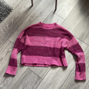 Rosa och vinröd/mörkrosa randig stickad tröja - Säljer en vacker stickad tröja med breda ränder i rosa och vinrött/mörkrosa. Tröjan har lång ärm, rund halsringning och en avslappnad passform. Perfekt för dig som gillar färg och vill ha något mysigt och snyggt! 