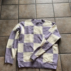 Lila och vit rutig stickad tröja från Lindex - Säljer en stickad tröja från Lindex i storlek 170. Tröjan har ett rutigt mönster i lila och vitt, rund halsringning och långa ärmar. 