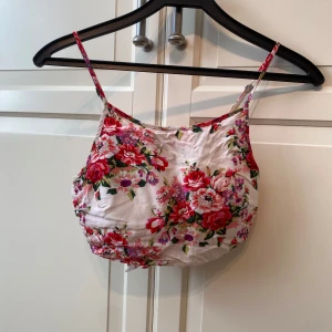 Blommig croppad linnetopp från Reverse - Supersöt croppad linnetopp från Reverse med blommigt mönster i rött, rosa och grönt på vit botten. Smala axelband, knytning i ryggen för en snygg passform och  inbyggd bh. Perfekt för varma dagar och somriga outfits!