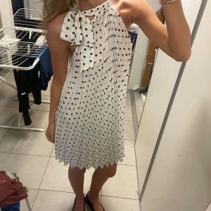 Vit prickig halterneckklänning med rosett - Säljer en söt vit kortklänning med svarta prickar och halterneck. Klänningen har en stor rosett vid halsen och ett lätt plisserat tyg! Superfin aldrig använd 