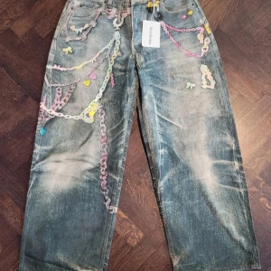 Unika jeansbyxor från Acne Studios med kedjedetaljer - Säljer ett par coola blå jeansbyxor från Acne Studios med färgglada kedjor och broderade detaljer. Byxorna har raka ben och en vintage tvättad look. Perfekta för dig som vill sticka ut med din stil!